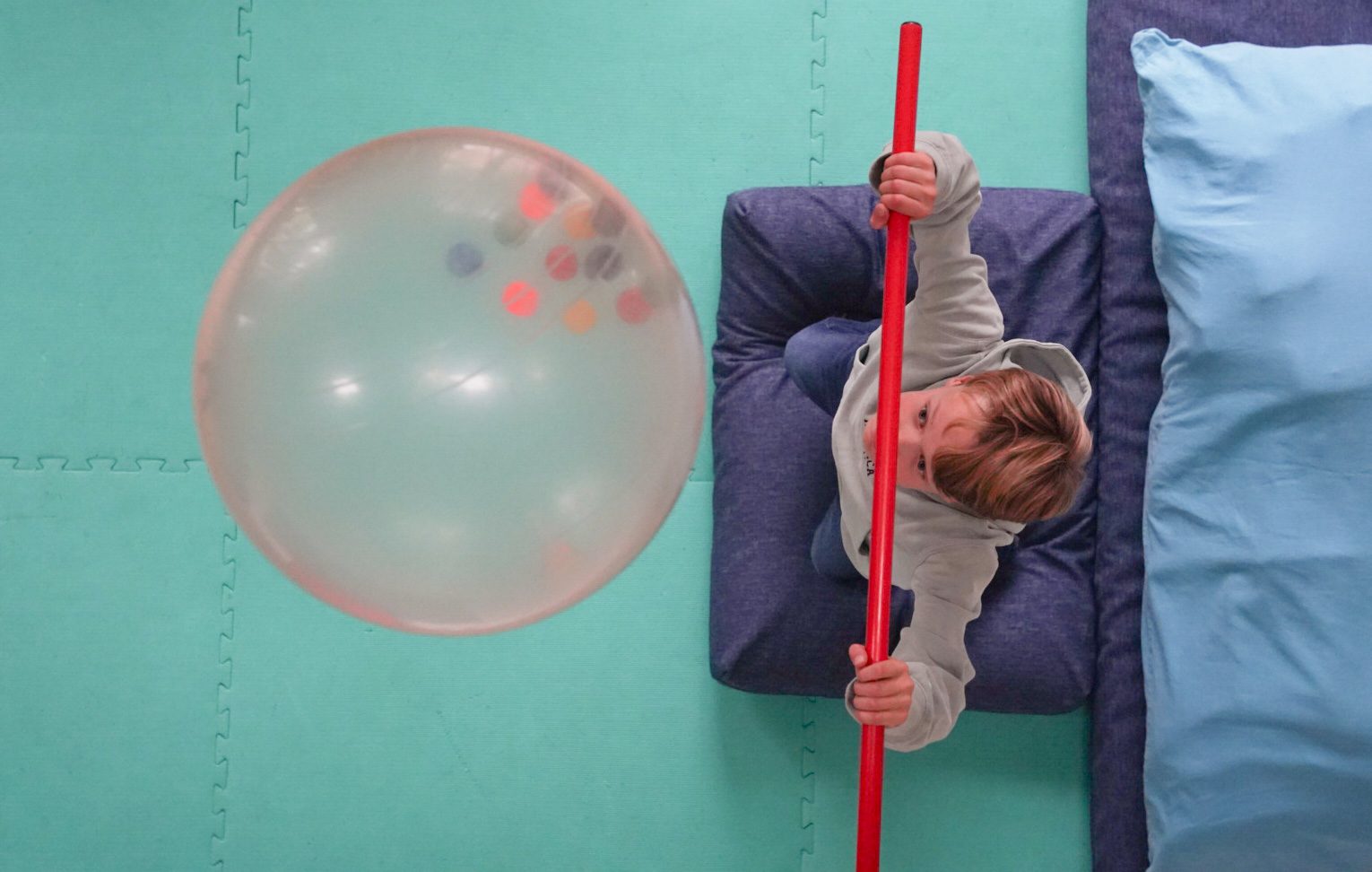 Een kind ligt op een kussen op de mat en slaat met een rode stok naar een grote ballon